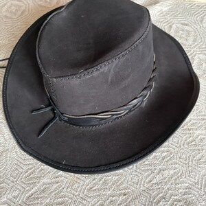 May include: A dark brown cowboy hat with a wide brim and a braided band. The hat has a structured crown and a leather tie. The hat is made of a suede-like material.