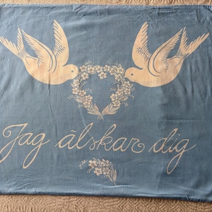 Puede incluir: Funda de almohada azul claro con dos palomas blancas enfrentadas, con un corazón floral entre ellas. La frase "Jag älskar dig" está escrita en blanco. Un pequeño diseño floral en la parte inferior.