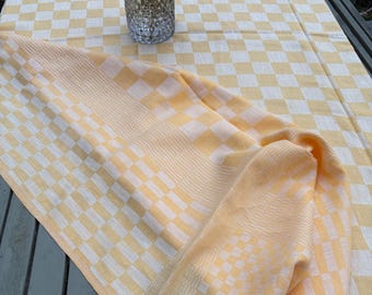 May include: A rectangular tablecloth featuring a yellow and white checkered pattern. A clear, textured glass vase containing a bouquet of yellow, purple, and green flowers sits on the surface of the fabric, which is draped over a wooden structure.