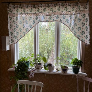 Puede incluir: Una cenefa de ventana de tela color crema con un patrón repetitivo de hojas de fresa verdes y pequeñas fresas rojas, acentuada por una banda horizontal de encaje decorativo beige a lo largo de la sección inferior de la cortina.