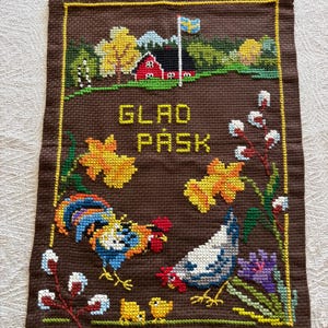 May include: Brown cross-stitch wall hanging with a yellow border. The design features a Swedish flag, a red house, yellow daffodils, and the words "GLAD PÅSK". A rooster, hen, chicks, and pussy willows are also depicted.