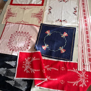 Könnte beinhalten: Eine Sammlung verschiedener weihnachtlicher Textilien, darunter bestickte Läufer, quadratische Tücher und Servietten. Zu den Motiven gehören rot-weiße Muster, ein blaues Tuch mit Engeln, ein schwarzes Tuch mit Wichteln und ein Läufer mit dem Text GOD JUL.