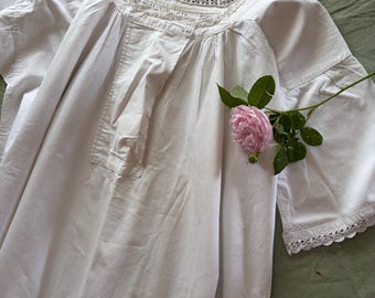 May include: A white cotton nightgown featuring a square neckline with delicate white lace trim, three-quarter length sleeves with matching lace cuffs, and a central vertical placket. A single light pink rose with green leaves rests on the fabric.