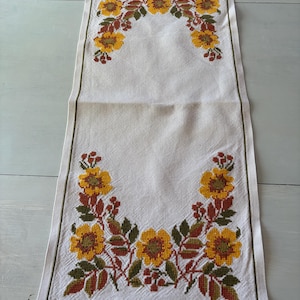 May include: Cream-coloured embroidered table runner with a floral design. The embroidery features yellow flowers, green leaves, and red berries. The runner has a green border and is made of a textured fabric.