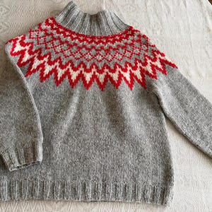 May include: A grey knitted jumper with a high neck and long sleeves. The yoke has a geometric pattern in red and white. The jumper is made of textured yarn and has ribbed cuffs and hem.