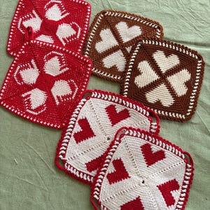 Op de afbeelding: Een set van vijf vierkante gehaakte pannenlappen met hartpatronen. Twee zijn rood met witte harten, twee zijn bruin met crèmekleurige harten en één is wit met rode harten. Elke pannenlap is voorzien van een klein lusje om op te hangen.