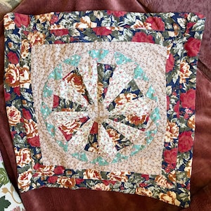 Pode incluir: Uma capa de almofada quadrada acolchoada com um padrão central de leque circular feito de vários tecidos florais em tons de vermelho, azul, verde e creme. O design central é rodeado por uma borda floral bege claro e uma moldura floral escura.