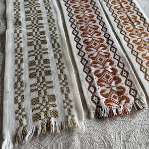 Puede incluir: Primer plano de varias tiras textiles tejidas con intrincados patrones geométricos en tonos marrón y crema. Cada tira tiene un borde con flecos. Los textiles parecen hechos a mano, mostrando una estética rústica y vintage.