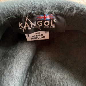 May include: A teal Kangol beret with a fuzzy texture. The beret has a black tag with the Kangol logo, the words "Made in Great Britain", and a Union Jack flag. The tag also includes the style name "Jaine" and the word "Regular".