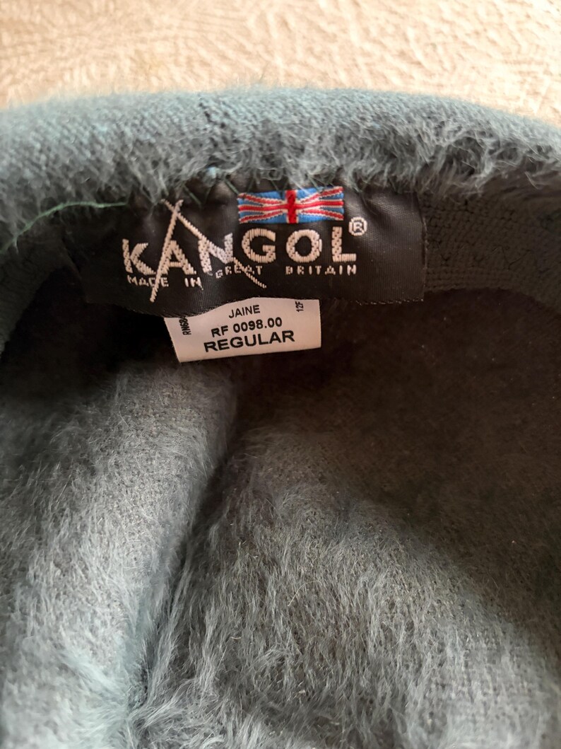 May include: A teal Kangol beret with a fuzzy texture. The beret has a black tag with the Kangol logo, the words "Made in Great Britain", and a Union Jack flag. The tag also includes the style name "Jaine" and the word "Regular".