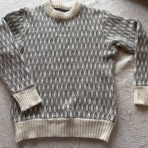 Könnte beinhalten: Ein Rundhals-Pullover mit einem geometrischen Muster in Schwarz und Off-White. Der Pullover hat gerippte Bündchen, Saum und Kragen in einem hellbeigen Farbton. Das Muster ist ein sich wiederholendes Design aus ineinandergreifenden Formen.