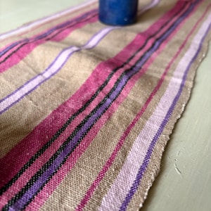Può includere: Runner da tavolo beige a righe verticali rosa, viola e nere. Il runner ha un bordo grezzo e una candela blu è posta sopra. Questo tessuto è adatto all'arredamento della casa.
