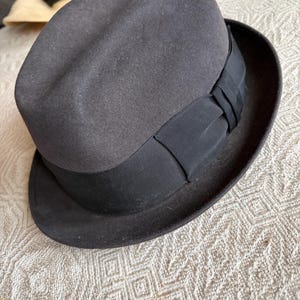 May include: A dark grey fedora hat with a black grosgrain ribbon band. The hat has a classic, structured shape with a slightly indented crown and a brim. The hat is made of a felt-like material.