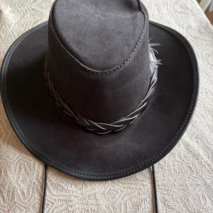 May include: A dark gray suede cowboy hat with a braided band and feather detail. The hat has a wide brim and a structured crown. A black chin strap is attached. This hat is suitable for outdoor activities.