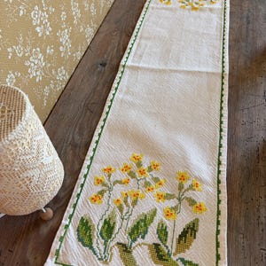 May include: Cream-coloured embroidered table runner with a green border. The runner features yellow and orange floral cross-stitch designs at each end. The flowers have green leaves and stems. The runner is displayed on a wooden table.