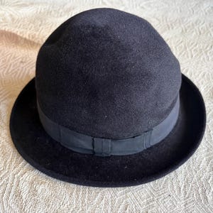 Peut inclure: Un chapeau melon noir avec une couronne arrondie et un large bord. Le chapeau est orné d'un ruban foncé à la base de la couronne. Le chapeau est fait d'une matière douce semblable à du feutre.