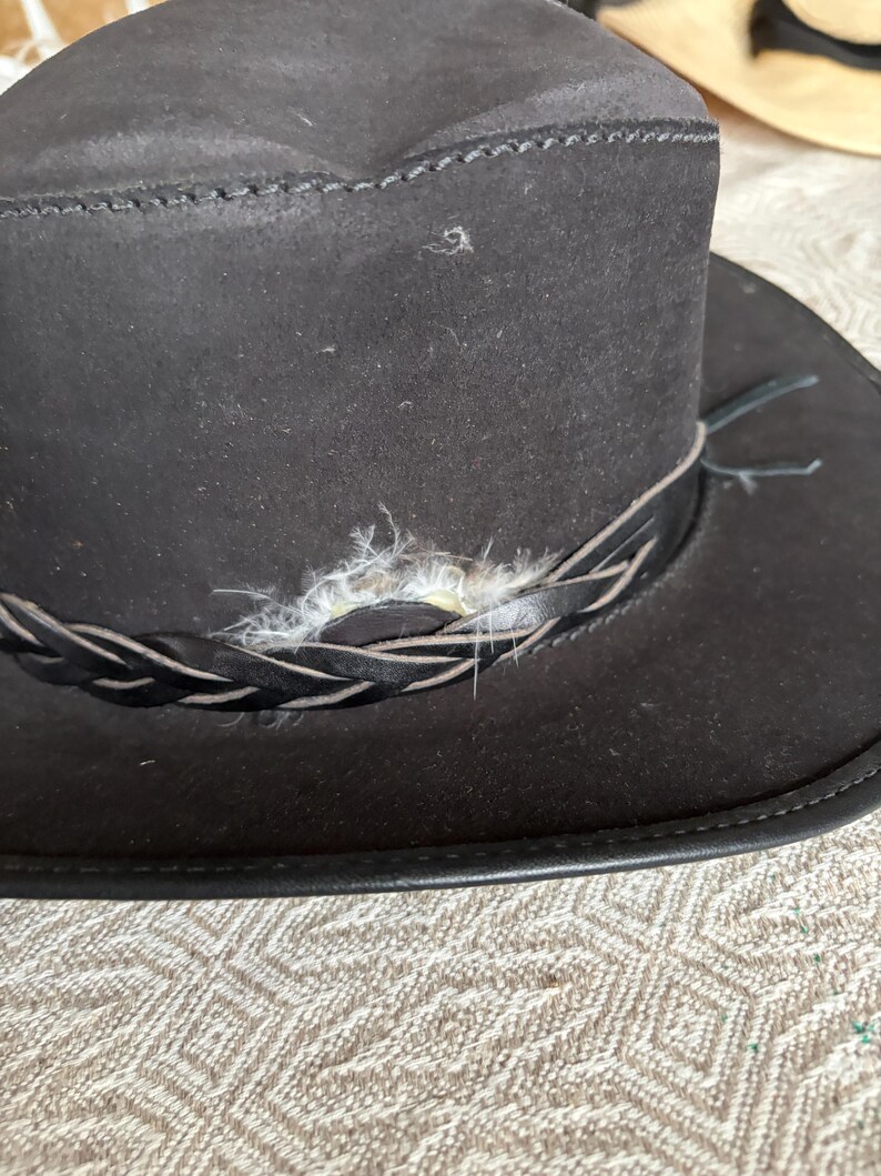 May include: A dark brown suede hat with a braided leather band and decorative feather accents. The hat has a wide brim and a structured crown, suitable for outdoor wear.
