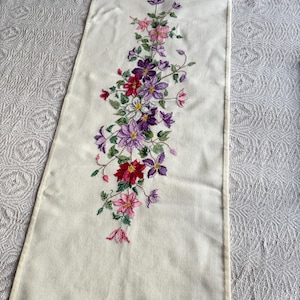 May include: Cream-coloured rectangular table runner with an embroidered floral design. The embroidery features a variety of flowers in shades of pink, purple, and red, with green leaves and stems. The runner is placed on a textured white surface.