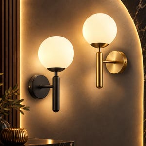 Könnte beinhalten: Zwei Wandleuchten mit kugelförmigen weißen Glasschirmen auf zylindrischen Metallarmen. Eine Leuchte hat ein mattschwarzes Finish, die andere ein gebürstetes Gold-Finish. Beide Lampen strahlen ein warmes, sanftes Licht an die Wandfläche ab.