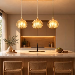 Peut inclure: Un luminaire suspendu à trois lampes avec un support de plafond rectangulaire en bois, trois globes en verre entourés de cages en métal doré, suspendus par des cordons blancs, diffusant une lumière chaude au-dessus d'un îlot de cuisine.