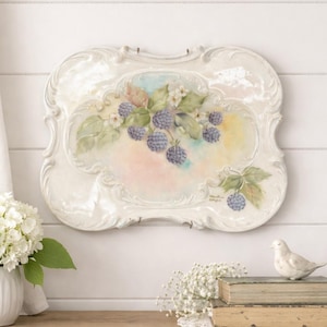 May include: A white, ornate, rectangular wall plaque with a hand-painted design of blackberries and leaves. The artwork features a pastel background with pink, blue, and yellow hues. The plaque has a glossy finish and is mounted on a white wall.