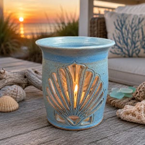 May include: A light blue ceramic candle holder with a seashell design cut-out. The candle holder is cylindrical with a flared top and a lit candle inside. The background shows a sunset over the ocean and beach-themed decor.