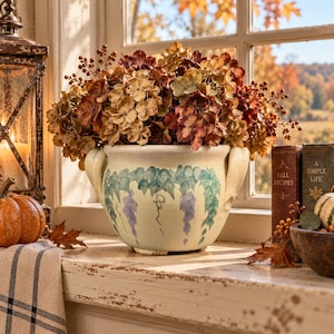 May include: A cream-colored ceramic planter with two side handles, featuring a hand-painted green ivy vine pattern and purple floral accents. The planter is filled with a dense arrangement of dried hydrangea blossoms in shades of brown and rust.