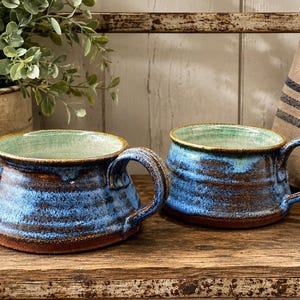 May include: Two wide-rimmed ceramic mugs featuring a reactive glaze with deep blue and brown tones. The interior of each mug is finished in a light, speckled mint green glaze, while the exterior displays a textured, earthy brown base.