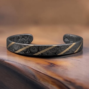 May include: A cuff bracelet crafted from dark gray Damascus steel featuring a swirling, wood-grain pattern throughout the metal. The band is accented with three thin, diagonal gold-colored metal stripes that cut across the textured surface.