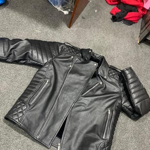 Pode incluir: Casaco de couro preto com detalhes acolchoados nos ombros e cotovelos. O casaco apresenta múltiplos bolsos com zíper e um fecho de zíper diagonal. O casaco está aberto, mostrando o forro interior.