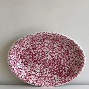 Op de afbeelding: Een ovale keramische serveerschaal met een witte basiskleur en een dicht, gevlekt patroon van diep magenta of donkerroze verfspatten die het gehele binnenoppervlak bedekken, wat zorgt voor een getextureerd en gespikkeld uiterlijk.