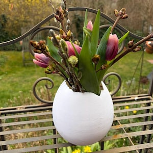 May include: A white, egg-shaped vase holds a bouquet of pink tulips and budding branches. The vase sits on a decorative metal shelf with a blurred outdoor background. The arrangement evokes a spring theme.