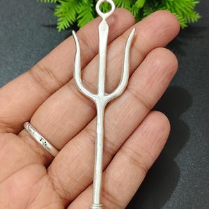 Puede incluir: Un colgante de tridente plateado con un lazo en la parte superior y tres puntas. El colgante se sostiene en una mano sobre un fondo oscuro con follaje verde. La parte inferior del colgante tiene un diseño texturizado.