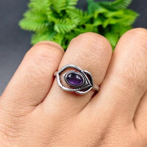 Puede incluir: Un anillo plateado con una piedra preciosa ovalada de color púrpura oscuro engastada en un diseño calado decorativo. El anillo es una pieza de joyería.