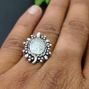 May include: A silver-colored ring featuring a turtle design. The turtle is in the center, surrounded by a beaded border. The ring is worn on a finger, showcasing the intricate details of the jewelry.