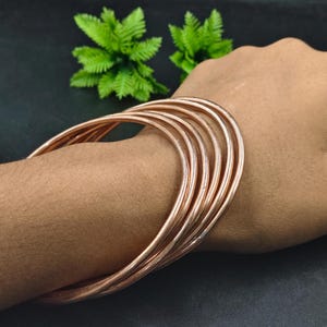 May include: A set of copper-colored bangles stacked on a wrist. The bangles are thin and round, with a smooth, metallic finish. The background is dark, with green foliage visible.