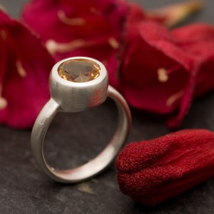 May include: A silver ring with a round, light brown gemstone set in a bezel setting. The ring is photographed on a dark gray surface with red flower petals.