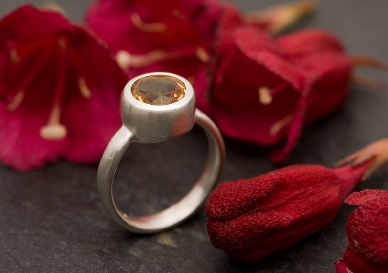 May include: A silver ring with a round, light brown gemstone set in a bezel setting. The ring is photographed on a dark gray surface with red flower petals.