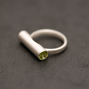 Pu&ograve; includere: Un anello d'argento con una pietra preziosa verde incastonata nella fascia. La fascia ha una forma semplice e cilindrica.