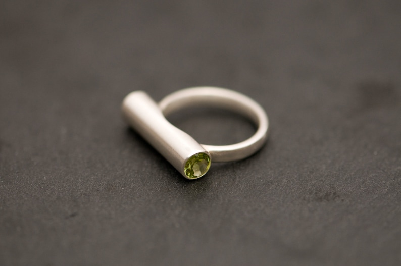 Pu&ograve; includere: Un anello d'argento con una pietra preziosa verde incastonata nella fascia. La fascia ha una forma semplice e cilindrica.