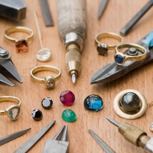 May include: A workbench with various jewelry making tools and gemstones. There are pliers, a small hammer, a screwdriver, a metal ruler, a gold chain, and a variety of gemstones in different colors and shapes.