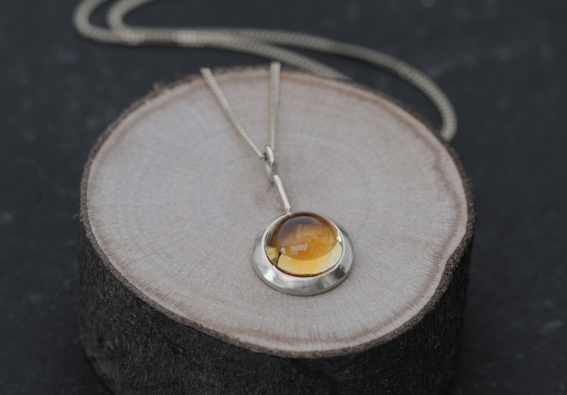 May include: A silver necklace with a round amber pendant. The pendant is set in a silver bezel and hangs from a delicate silver chain.