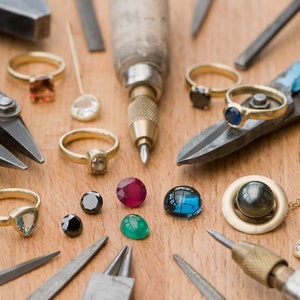 May include: A workbench with various jewelry making tools and gemstones. There are pliers, a small hammer, a screwdriver, a metal ruler, a gold chain, and a variety of gemstones in different colors and shapes.
