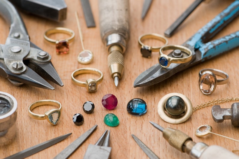 May include: A workbench with various jewelry making tools and gemstones. There are pliers, a small hammer, a screwdriver, a metal ruler, a gold chain, and a variety of gemstones in different colors and shapes.