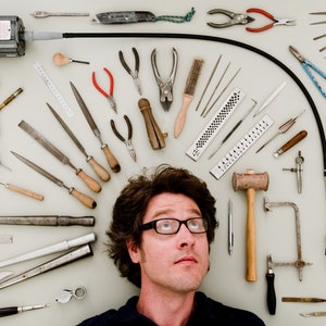 May include: A variety of tools and equipment are arranged in a circle around a person's head. The tools include files, pliers, a hammer, a saw, a drill, and a torch. The person is looking up at the camera.