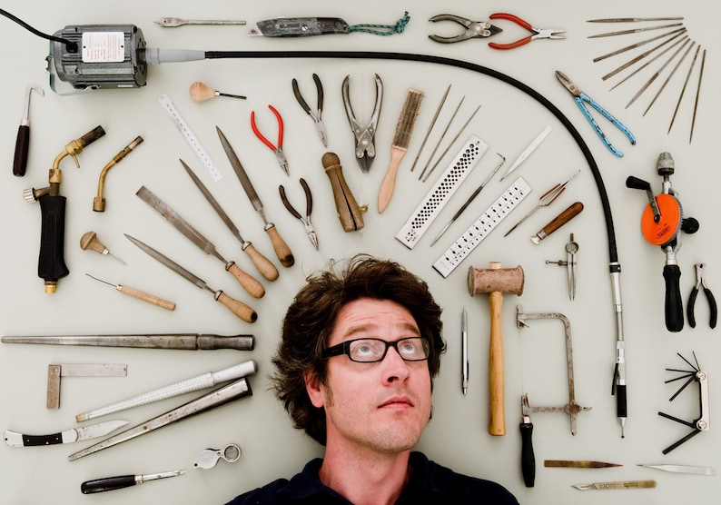 May include: A variety of tools and equipment are arranged in a circle around a person's head. The tools include files, pliers, a hammer, a saw, a drill, and a torch. The person is looking up at the camera.