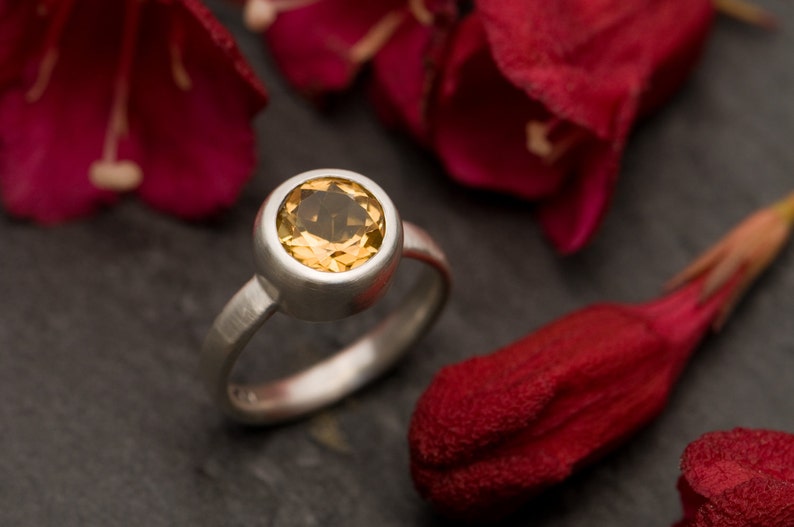 May include: A silver ring with a round, bezel-set, yellow gemstone. The ring is set against a background of red flower petals.