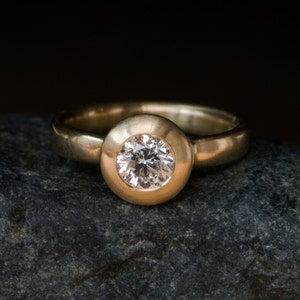 May include: A gold ring with a round, bezel-set diamond. The ring is sitting on a grey rock.