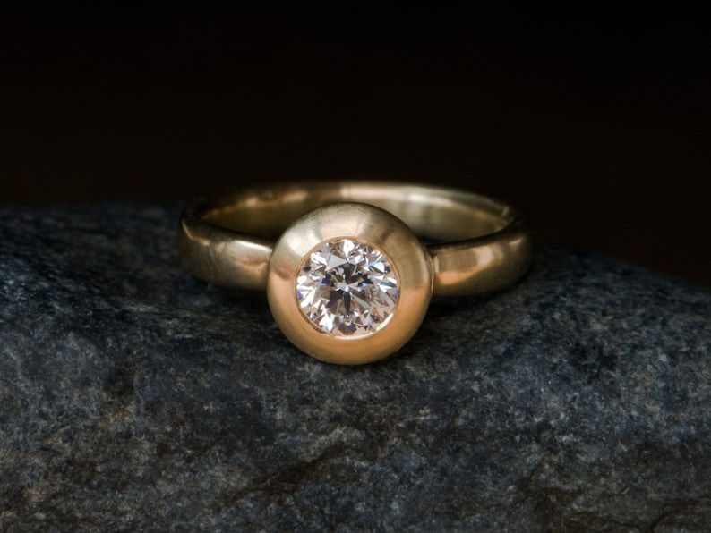 May include: A gold ring with a round, bezel-set diamond. The ring is sitting on a grey rock.
