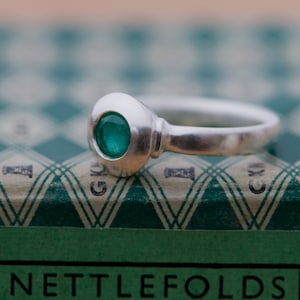May include: A silver ring with a green gemstone set in a round bezel. The ring is sitting on a green and white patterned box with the word "NETTLEFOLDS" printed on the front.